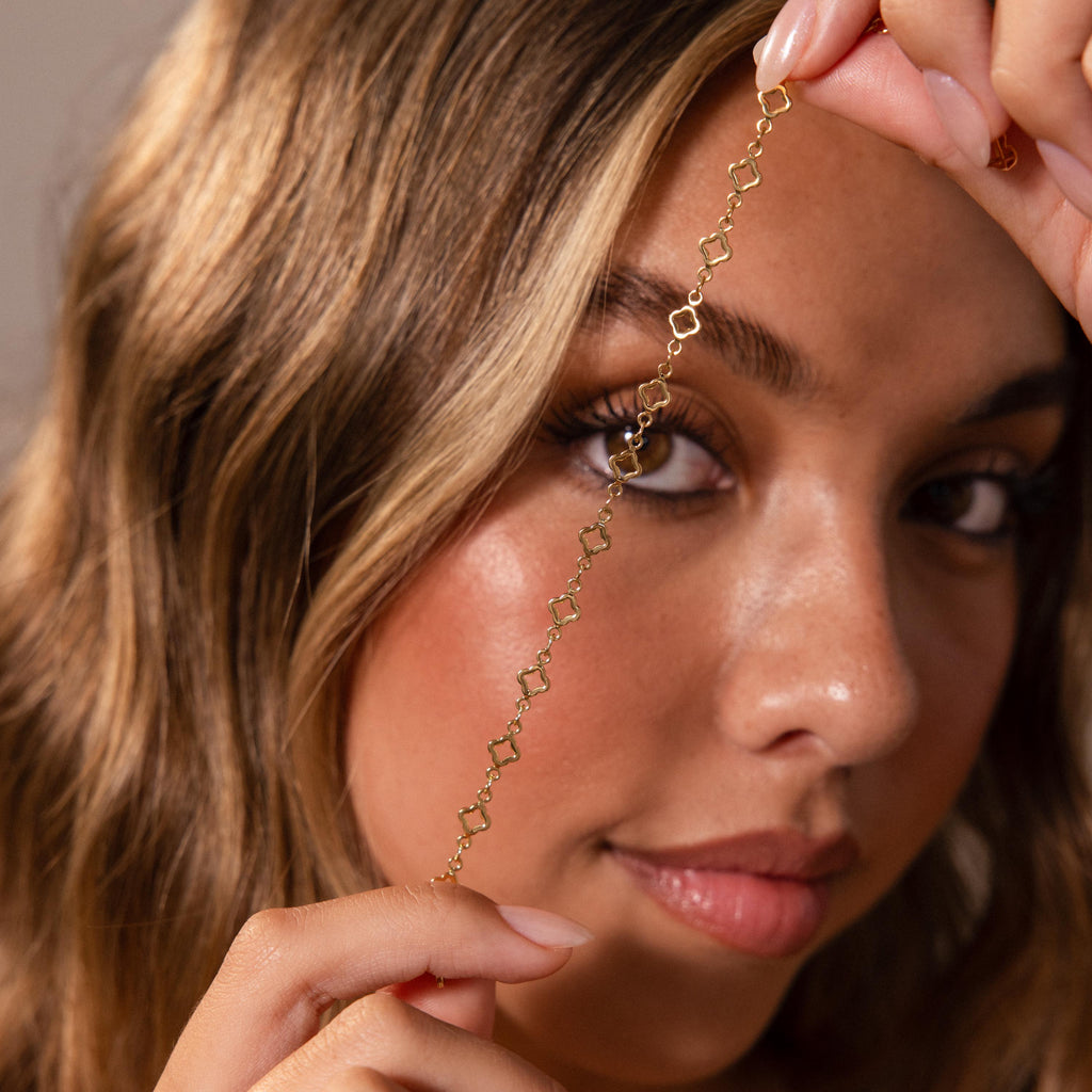 A woman holds a delicate gold chain with geometric links by her face, softly gazing at the camera while wearing the Clover Station Bracelet.