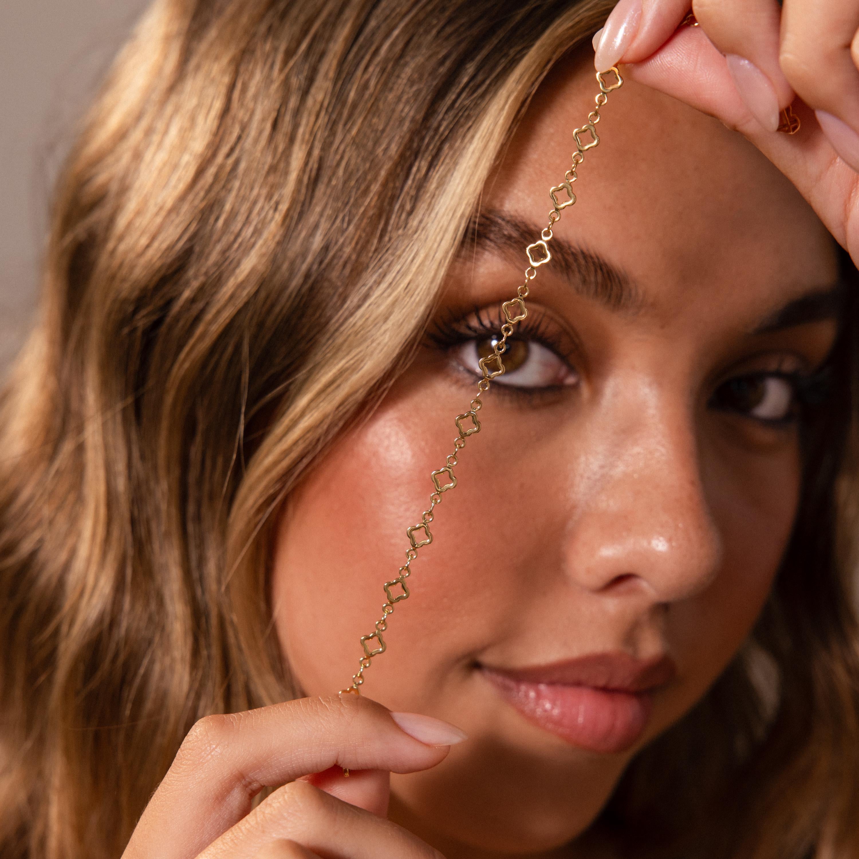 A woman holds a delicate gold chain with geometric links by her face, softly gazing at the camera while wearing the Clover Station Bracelet.