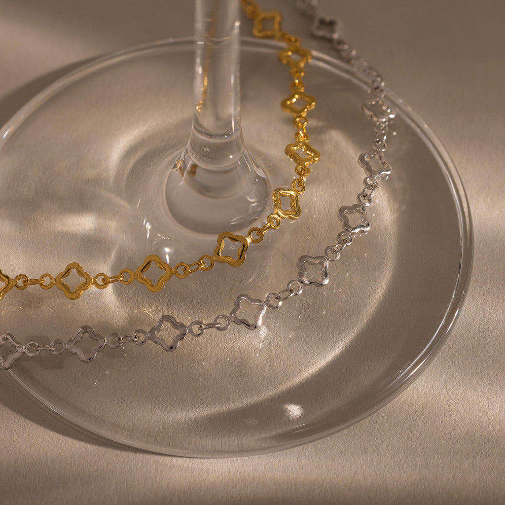 The Clover Station Bracelet, a geometric chain bracelet with nature-inspired details in gold and silver, is displayed around the base of a glass on a beige surface.
