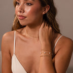 Woman in a cream dress wearing gold jewelry, including the Clover Station Bracelet in 18K Gold, looks to the side with her hand touching her neck.