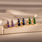 Three pairs of Ombre Topaz Flatback Studs in gold, with purple, blue, and green gemstones, create vibrant earrings—a thoughtful personalized gift displayed on a beige textured surface.