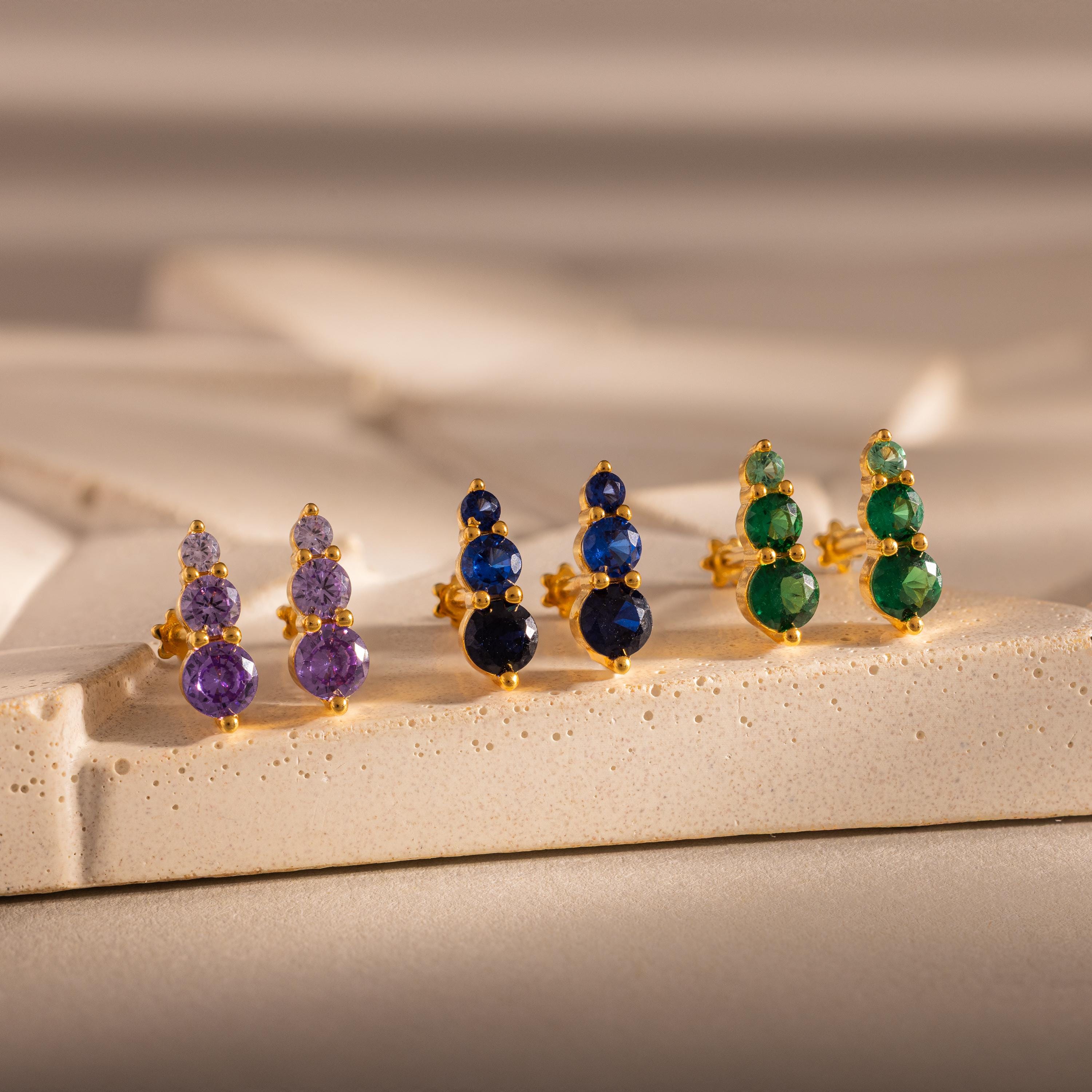 Three pairs of Ombre Topaz Flatback Studs in gold, with purple, blue, and green gemstones, create vibrant earrings—a thoughtful personalized gift displayed on a beige textured surface.