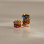 Six jeweled rings in various colors, including a Pave Garnet Charm Spacer, are stacked in two columns on a reflective, neutral surface.
