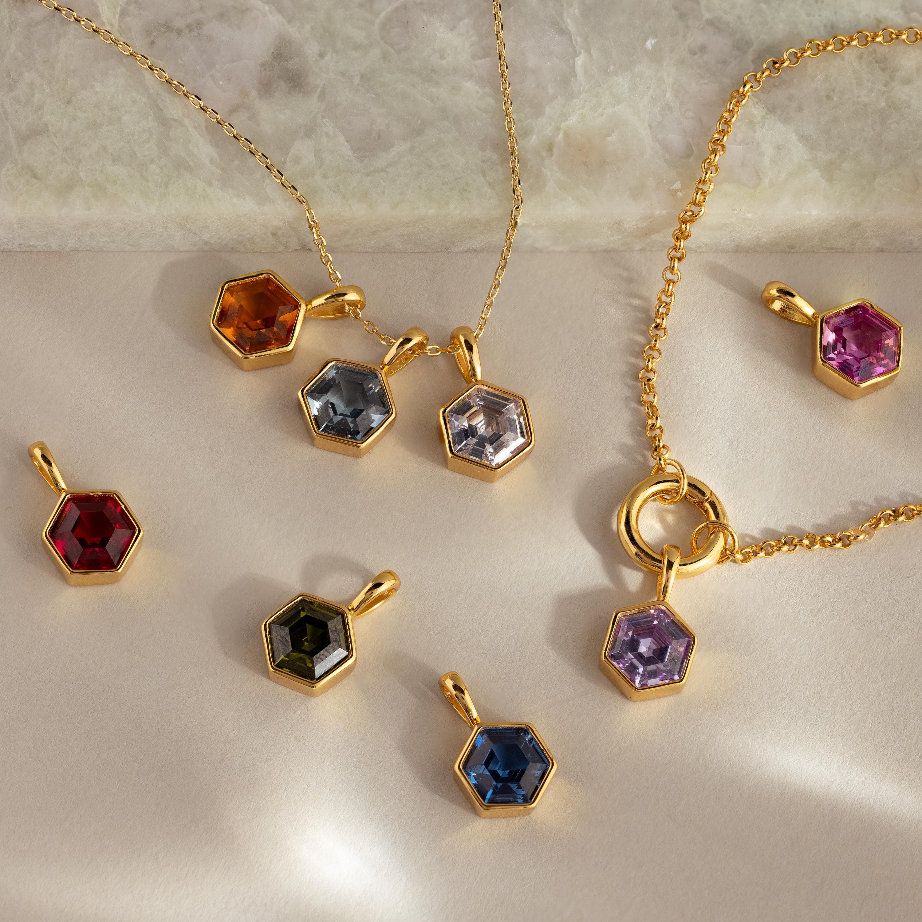 The Hexagon Blue Zircon Charm features a faceted blue zircon stone in a gold geometric frame, displayed on a marble and beige background.