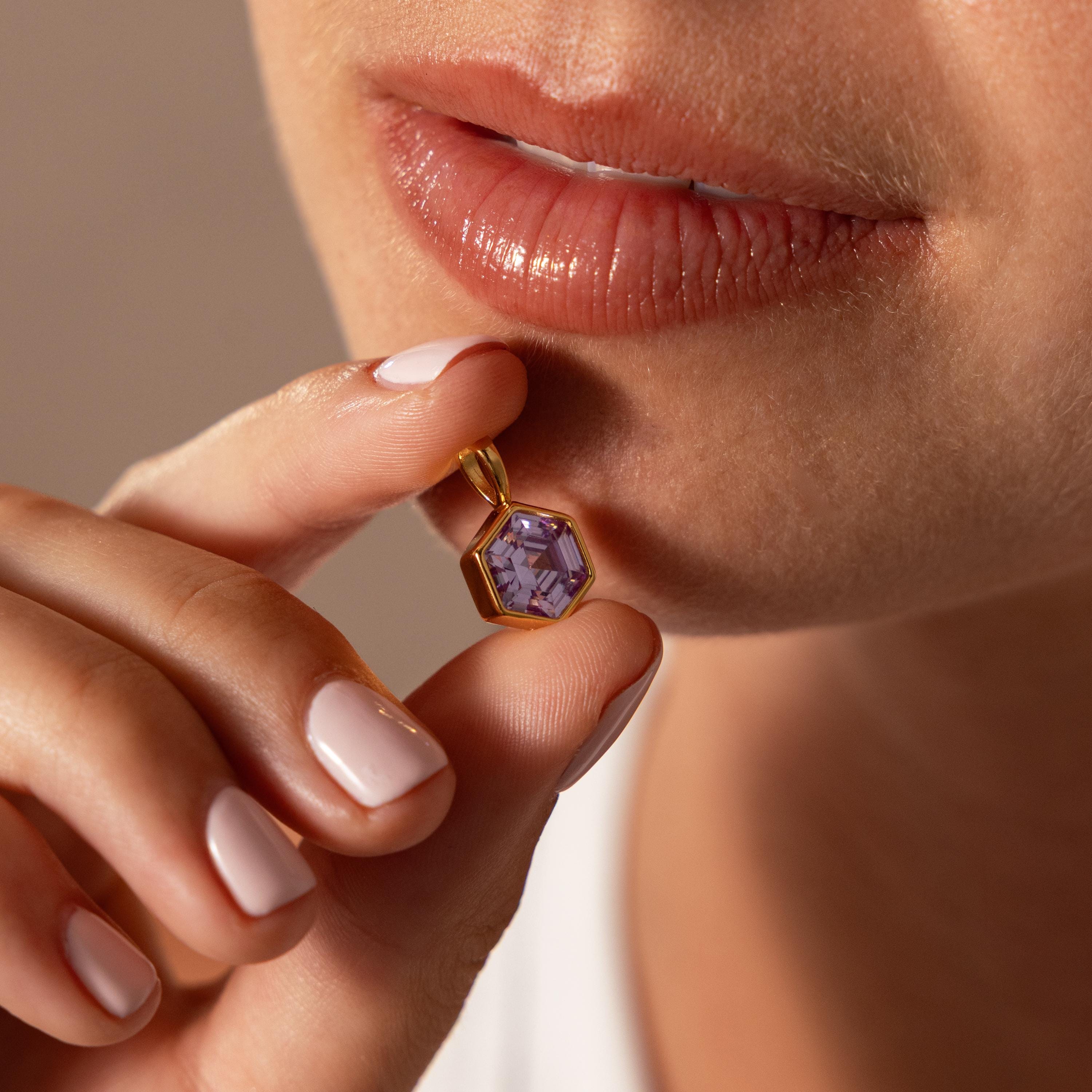 Someone holds the Hexagon Birthstone Charm featuring a faceted purple stone close to their lips.