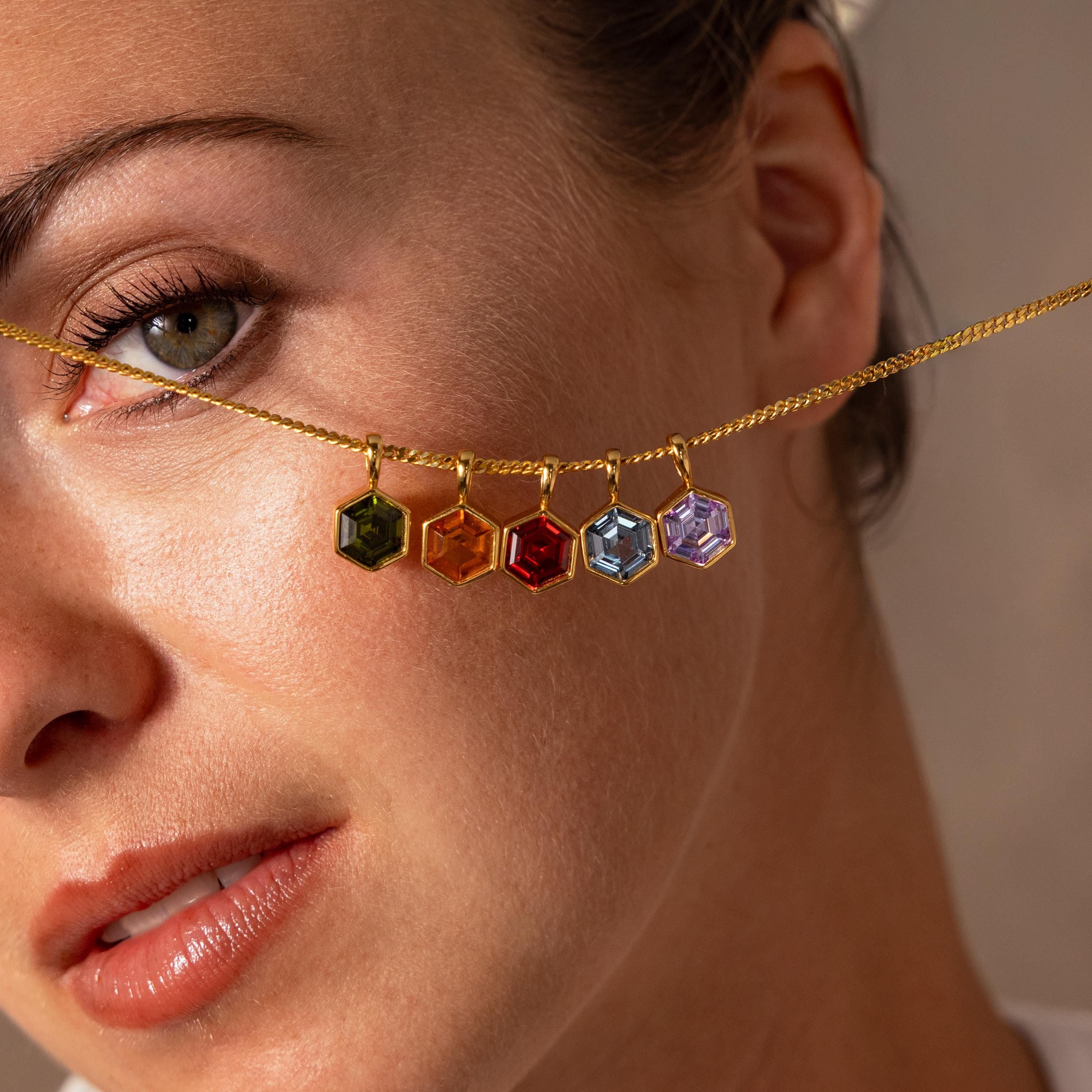 Woman holding a gold necklace with five colorful Hexagon Topaz Charms across her face, showcasing vibrant geometric jewelry.