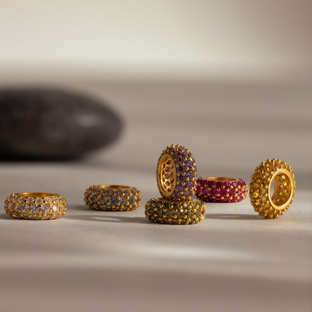 Six gold rings, some featuring Pavé Alexandrite Charm Spacers and vibrant birthstones, are arranged on a neutral surface with a blurred dark stone in the background.