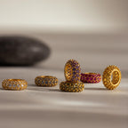 Six gold rings, some featuring Pavé Alexandrite Charm Spacers and vibrant birthstones, are arranged on a neutral surface with a blurred dark stone in the background.