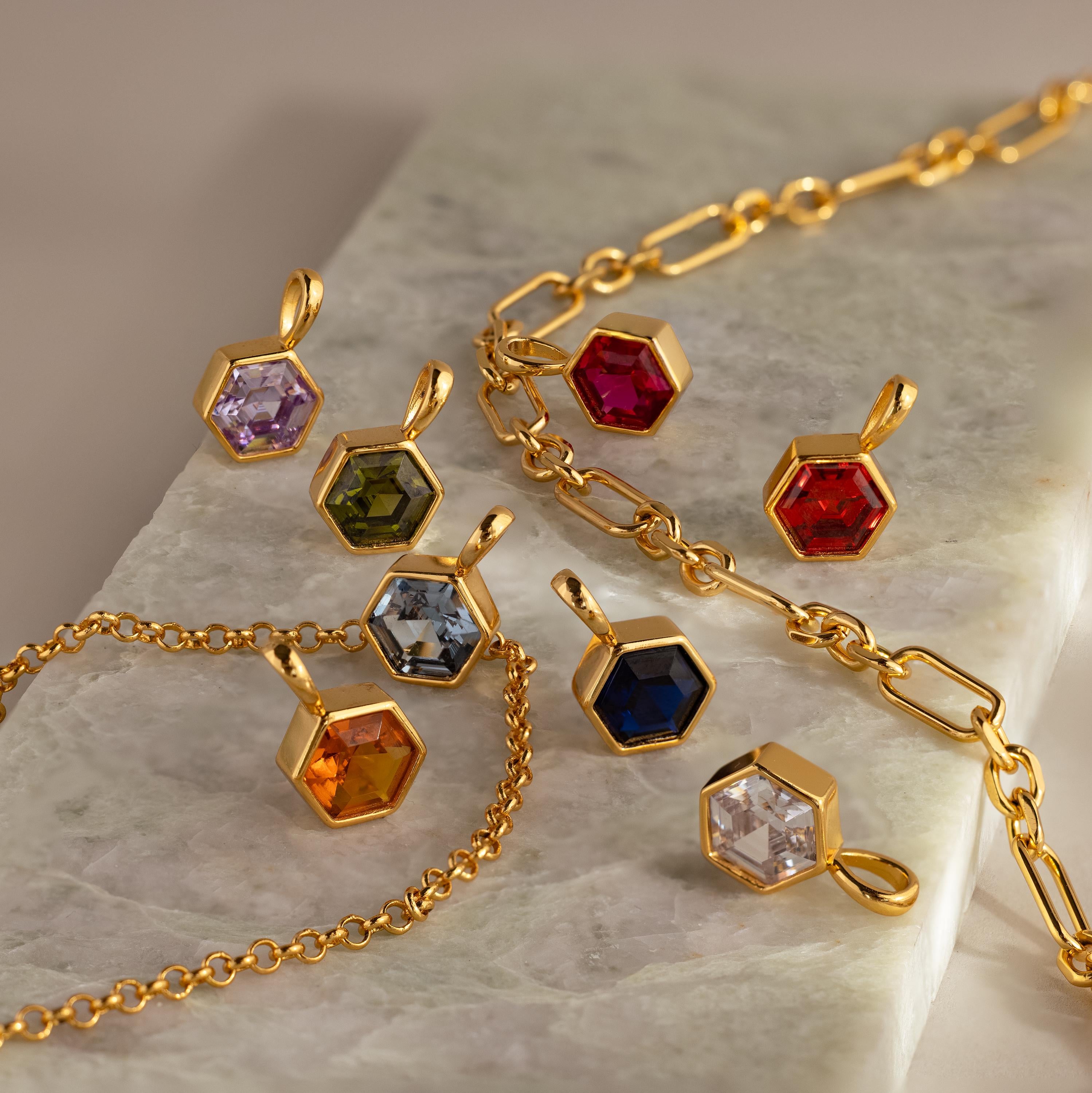 Elegant gold geometric jewelry featuring the Hexagon Topaz Charm with faceted stones in various colors, beautifully displayed on a marble surface.