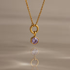 The Hexagon Birthstone Charm features a faceted stone in a hexagonal pendant, displayed on a gold necklace against a soft beige background with a reflection—a striking piece of geometric jewelry.