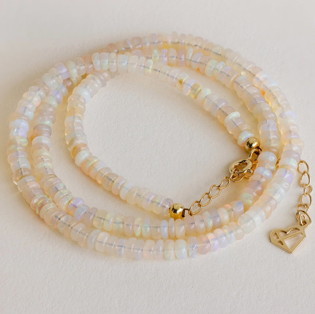 The Opal Beaded Necklace features iridescent beads, a gold clasp, and a heart-shaped charm—adding bohemian flair and shimmering beauty to any look.