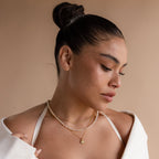 A woman with a slicked-back bun wears the Opal Beaded Necklace layered with gold chains and an off-shoulder white top, set against a beige background.