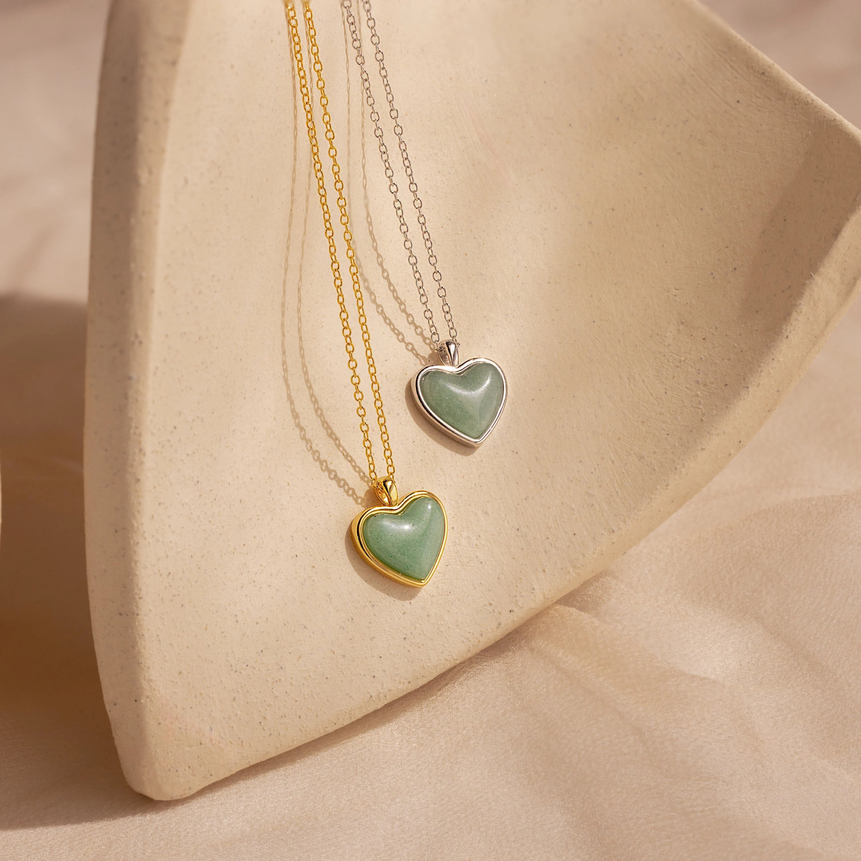 Two heart-shaped Aventurine Heart Necklaces—one gold and one silver—sparkle with aventurine gemstones on a textured beige surface. Ideal for lovers of this elegant necklace style.