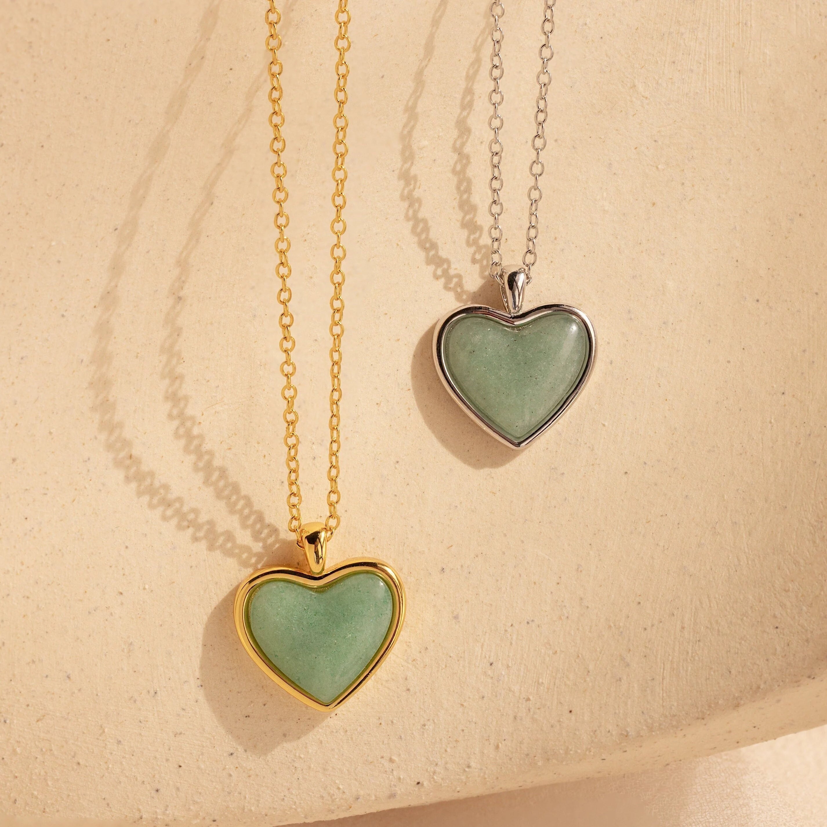 Two Aventurine Heart Necklaces—one on a gold chain, one on a silver chain—are displayed against a beige background.