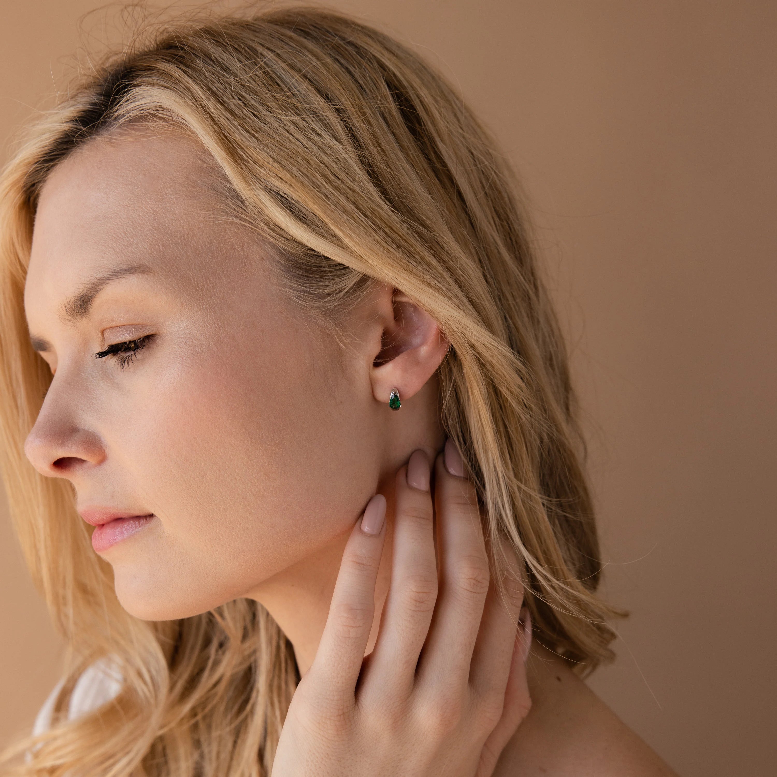 A blonde woman touches her neck while wearing Emerald Teardrop Huggies, pictured against a neutral background.