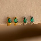 The Emerald Teardrop Huggies, available in gold or silver settings, feature dainty green stones that add elegance to any look. These stylish hoop earrings are shown beautifully displayed on a beige surface.