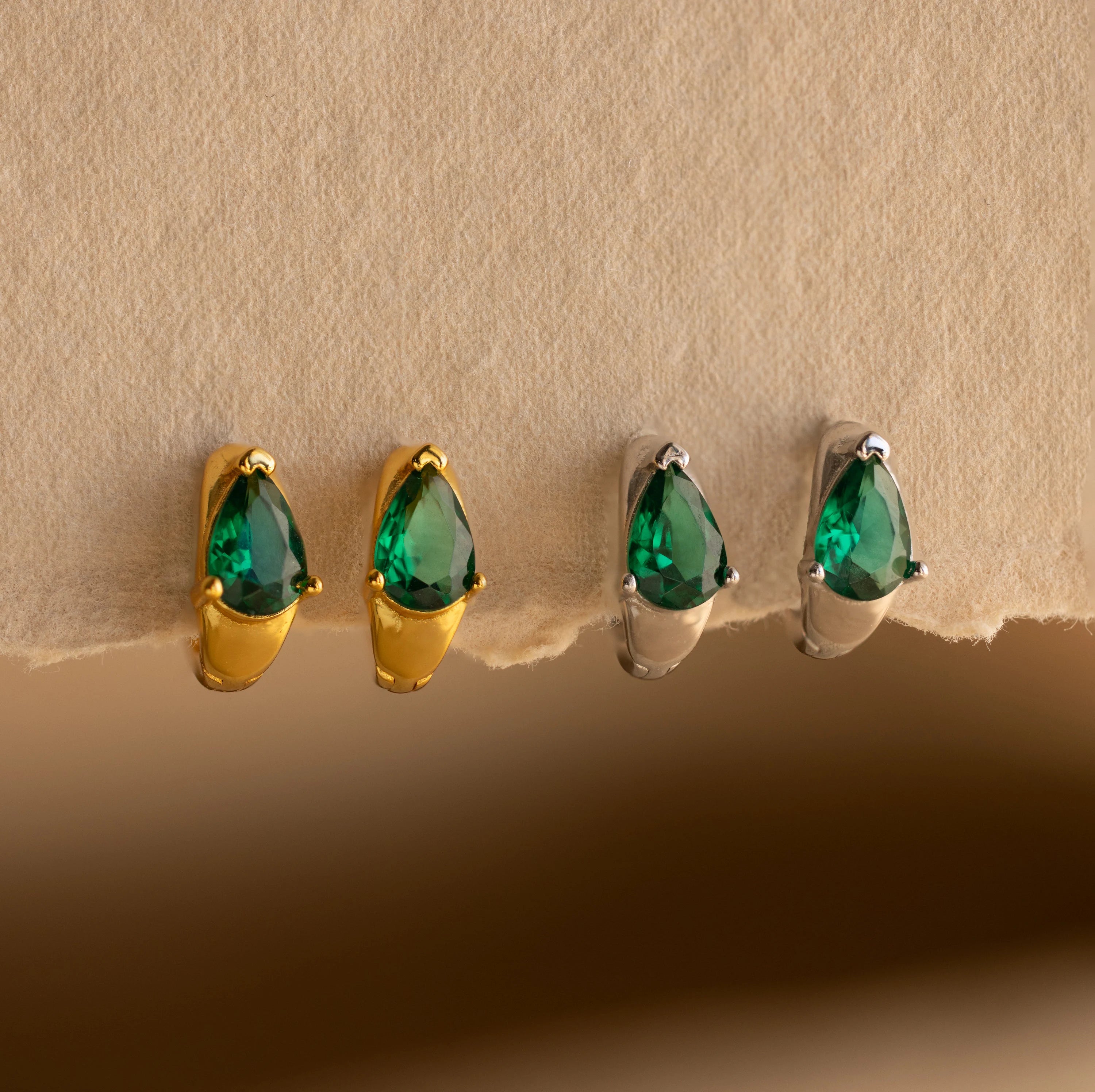 The Emerald Teardrop Huggies, available in gold or silver settings, feature dainty green stones that add elegance to any look. These stylish hoop earrings are shown beautifully displayed on a beige surface.