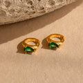 Emerald Teardrop Huggies—dainty hoop earrings with green gemstones—rest on a beige surface beside a textured stone, offering an elegant update to the classic style.