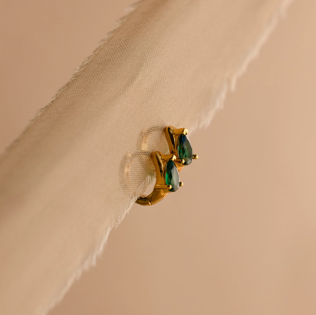 Emerald Teardrop Huggies shine with green gemstones, elegantly displayed on a sheer beige fabric background.