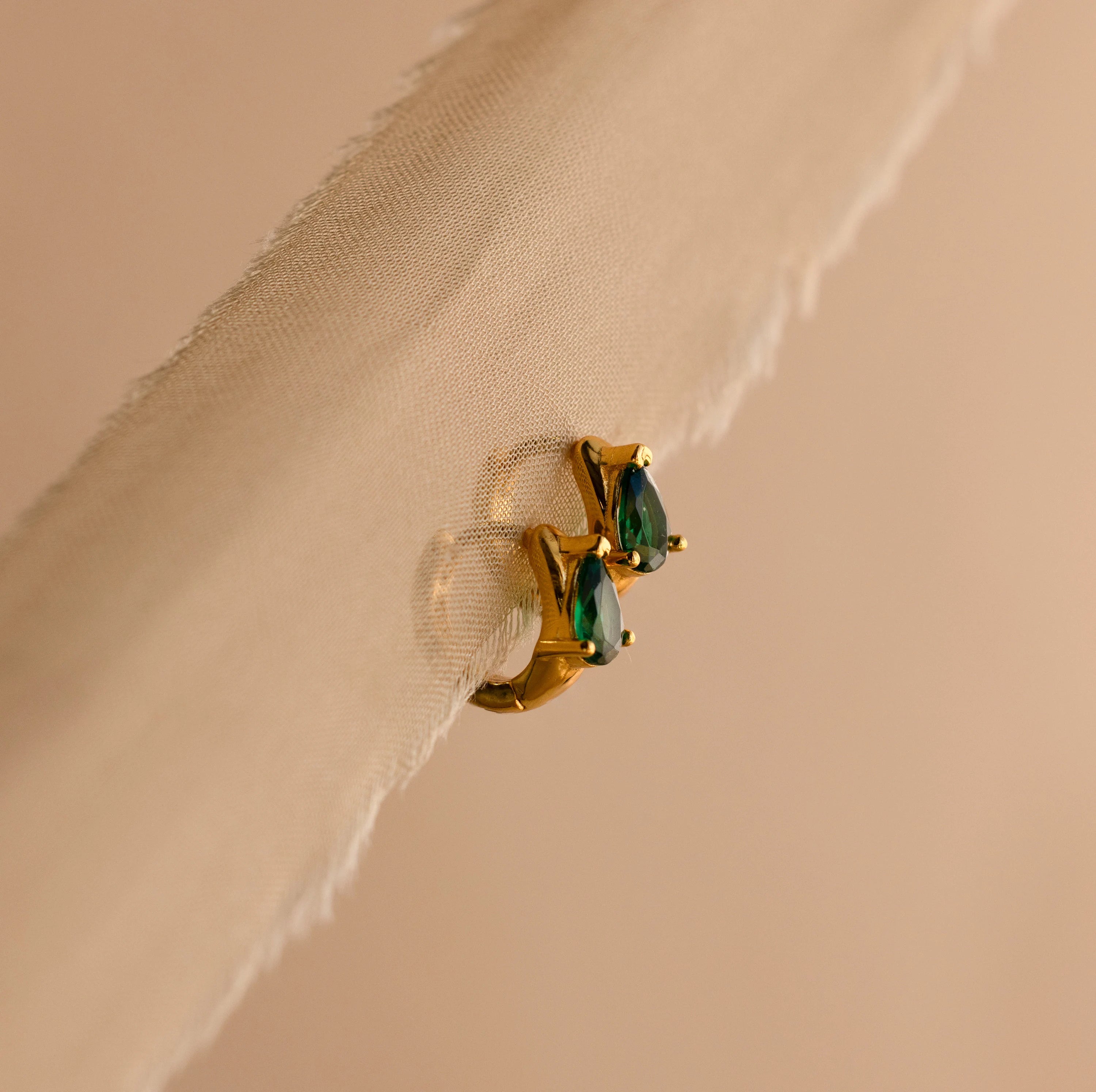 Emerald Teardrop Huggies shine with green gemstones, elegantly displayed on a sheer beige fabric background.