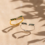 Two dainty Open Moss Agate Chevron Rings—one gold, one silver with green stones—rest on a beige surface. This charming set is perfect as promise rings.