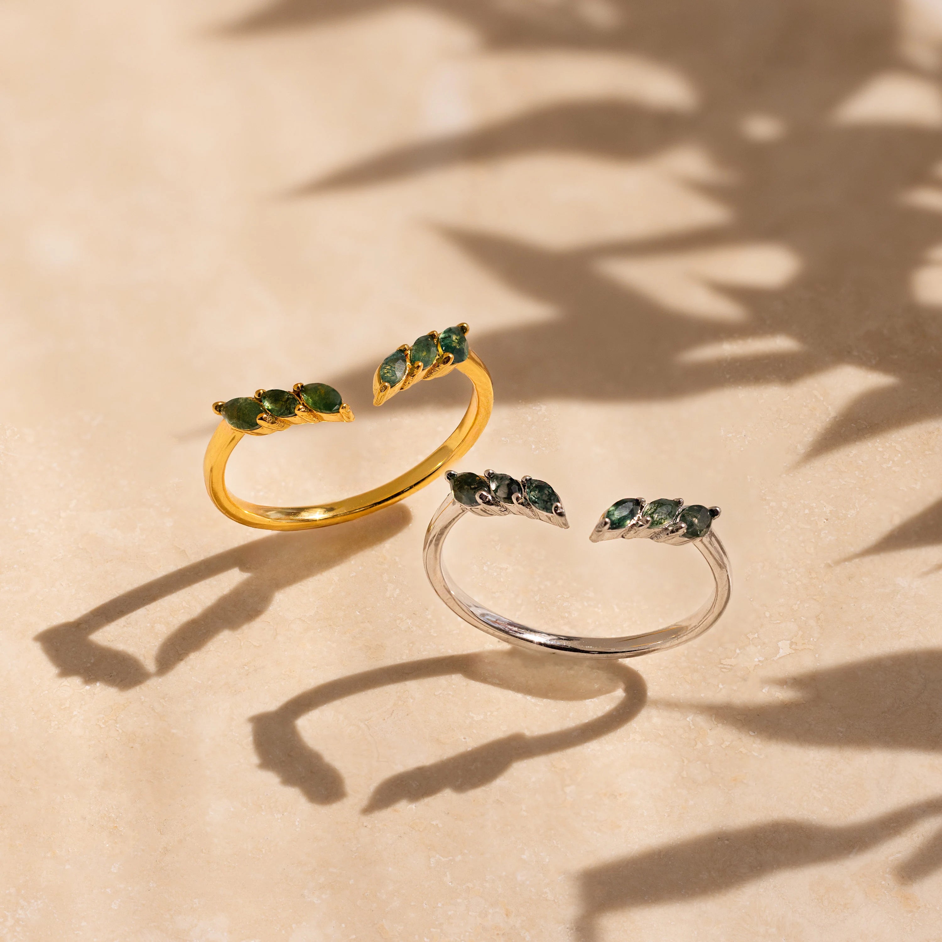 Two dainty Open Moss Agate Chevron Rings—one gold, one silver with green stones—rest on a beige surface. This charming set is perfect as promise rings.