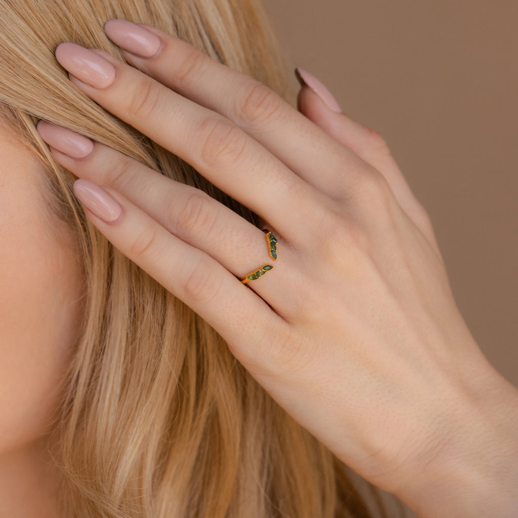 A hand with nude polished nails touches blonde hair, adorned with the Open Moss Agate Chevron Ring—a delicate gold band set with green stones.