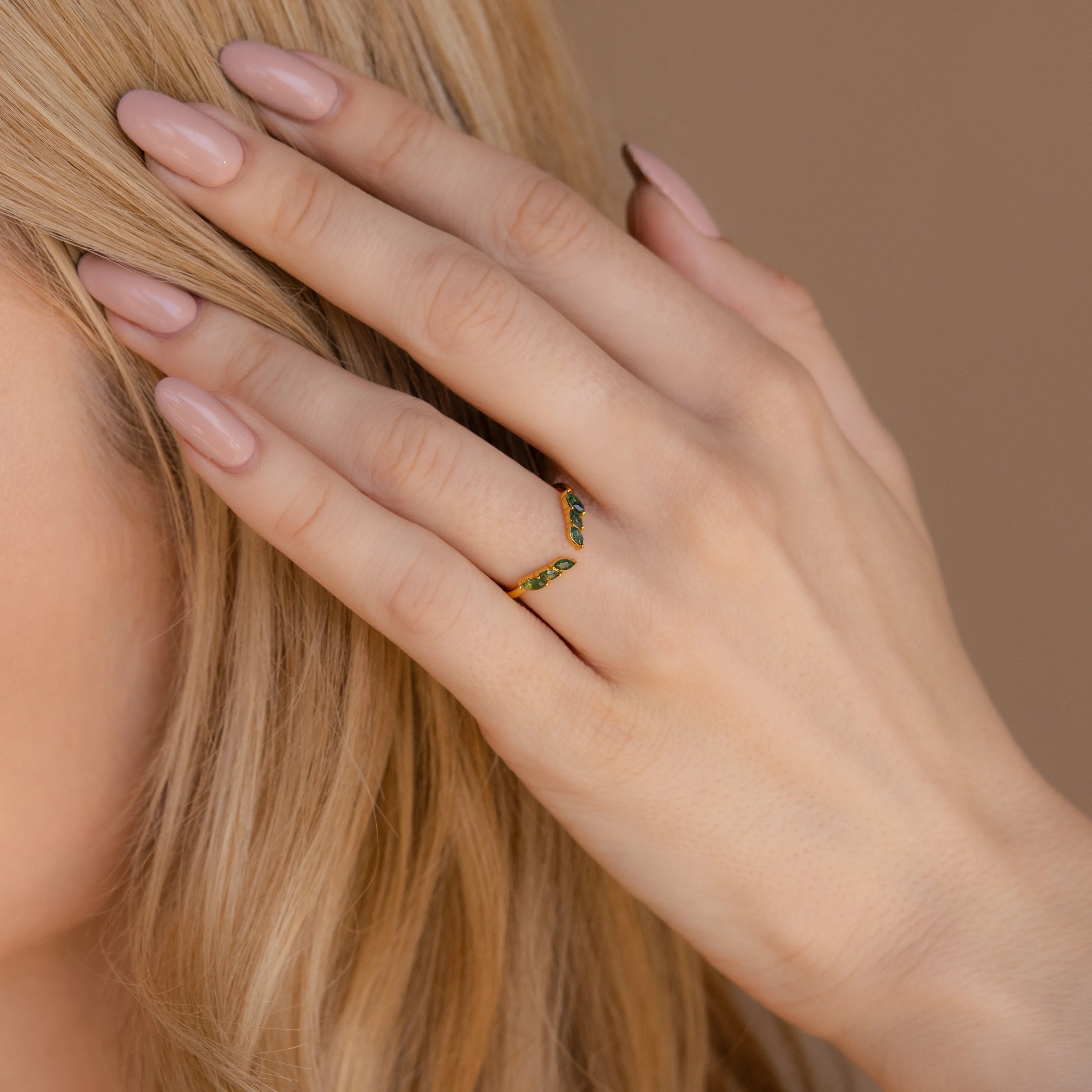 A hand with nude polished nails touches blonde hair, adorned with the Open Moss Agate Chevron Ring—a delicate gold band set with green stones.