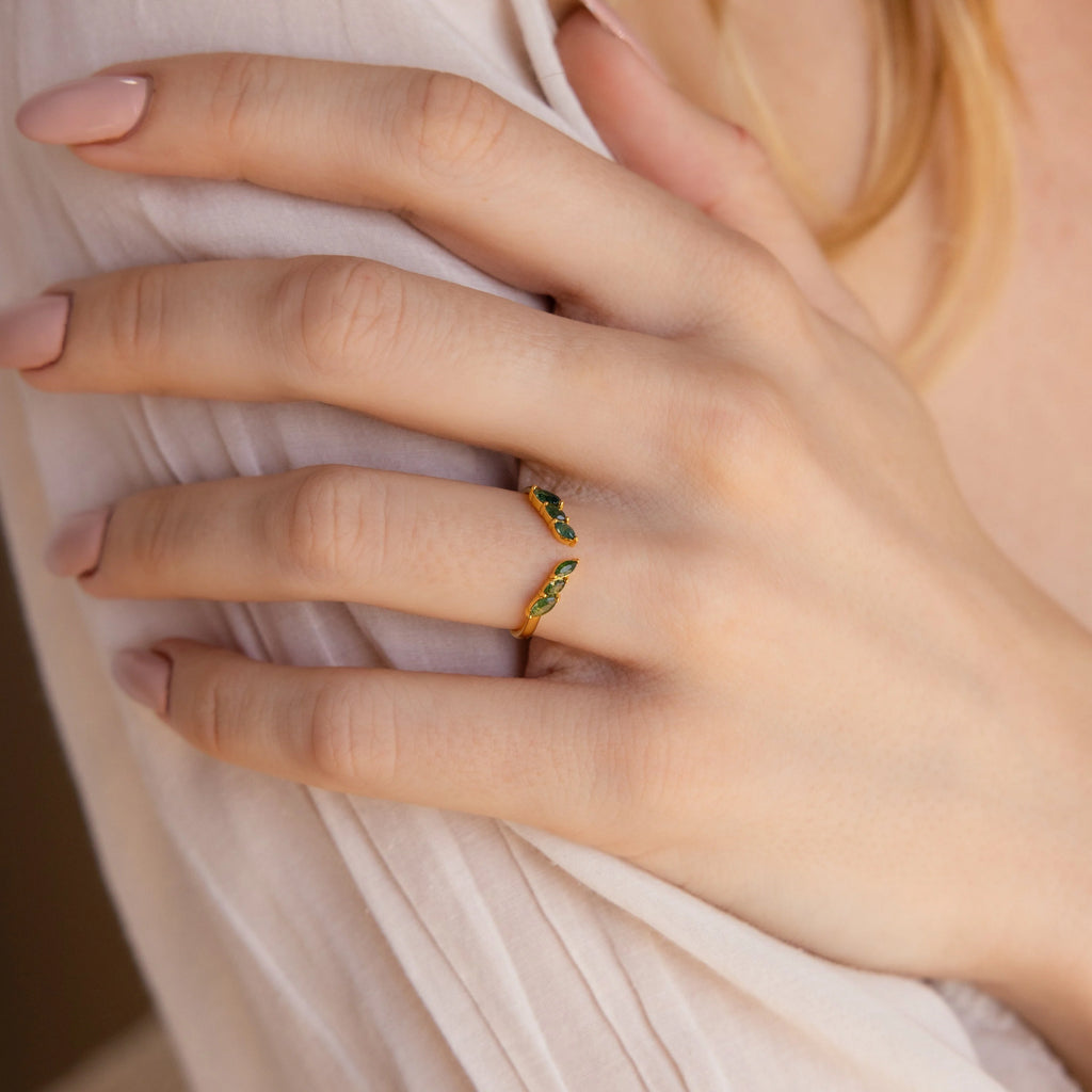 A manicured hand wears a dainty ring and the delicate Open Moss Agate Chevron Ring, both adorned with small green gemstones.