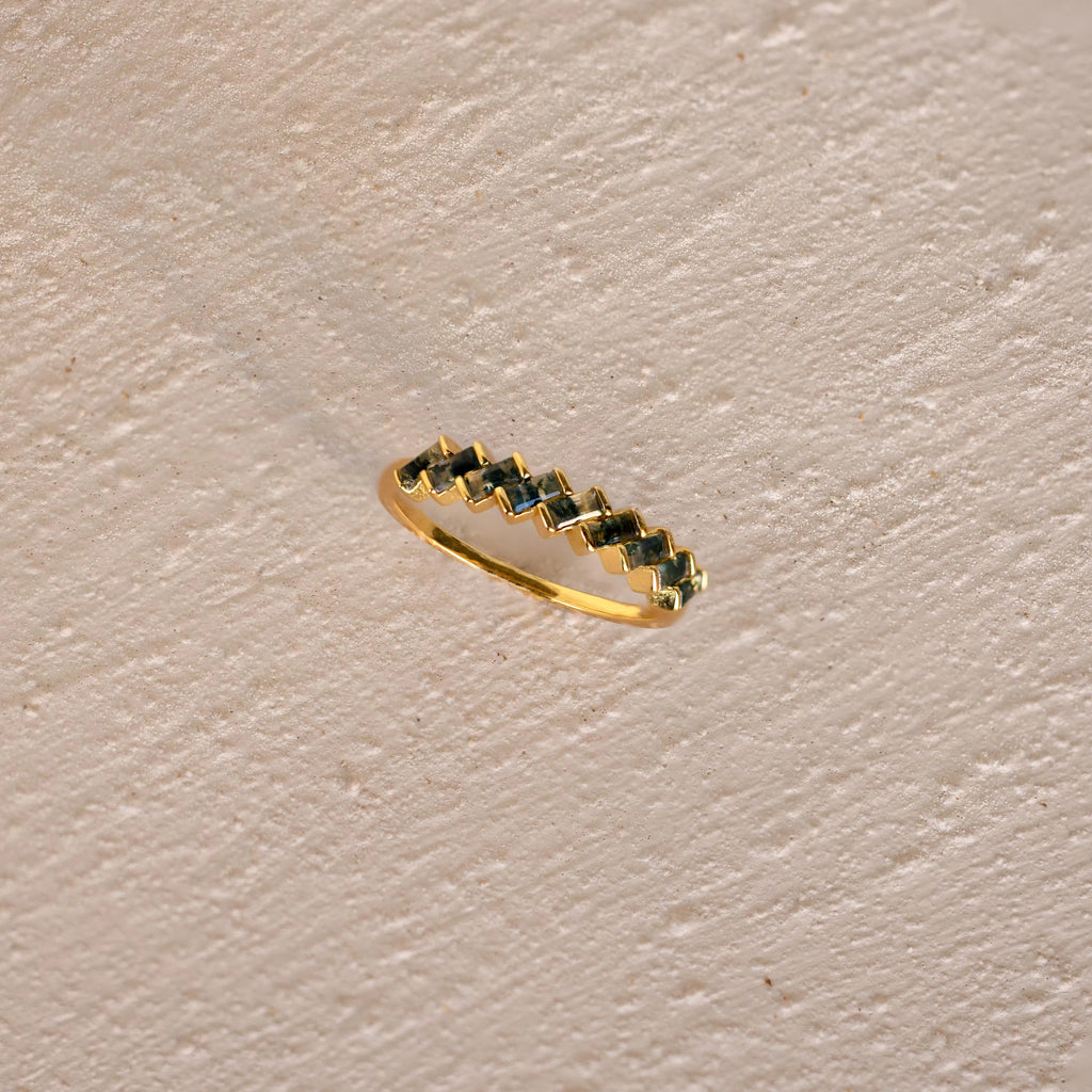 The Moss Agate Eternity Ring, featuring a nature-inspired zigzag pattern, rests on a textured beige surface, embodying organic elegance.