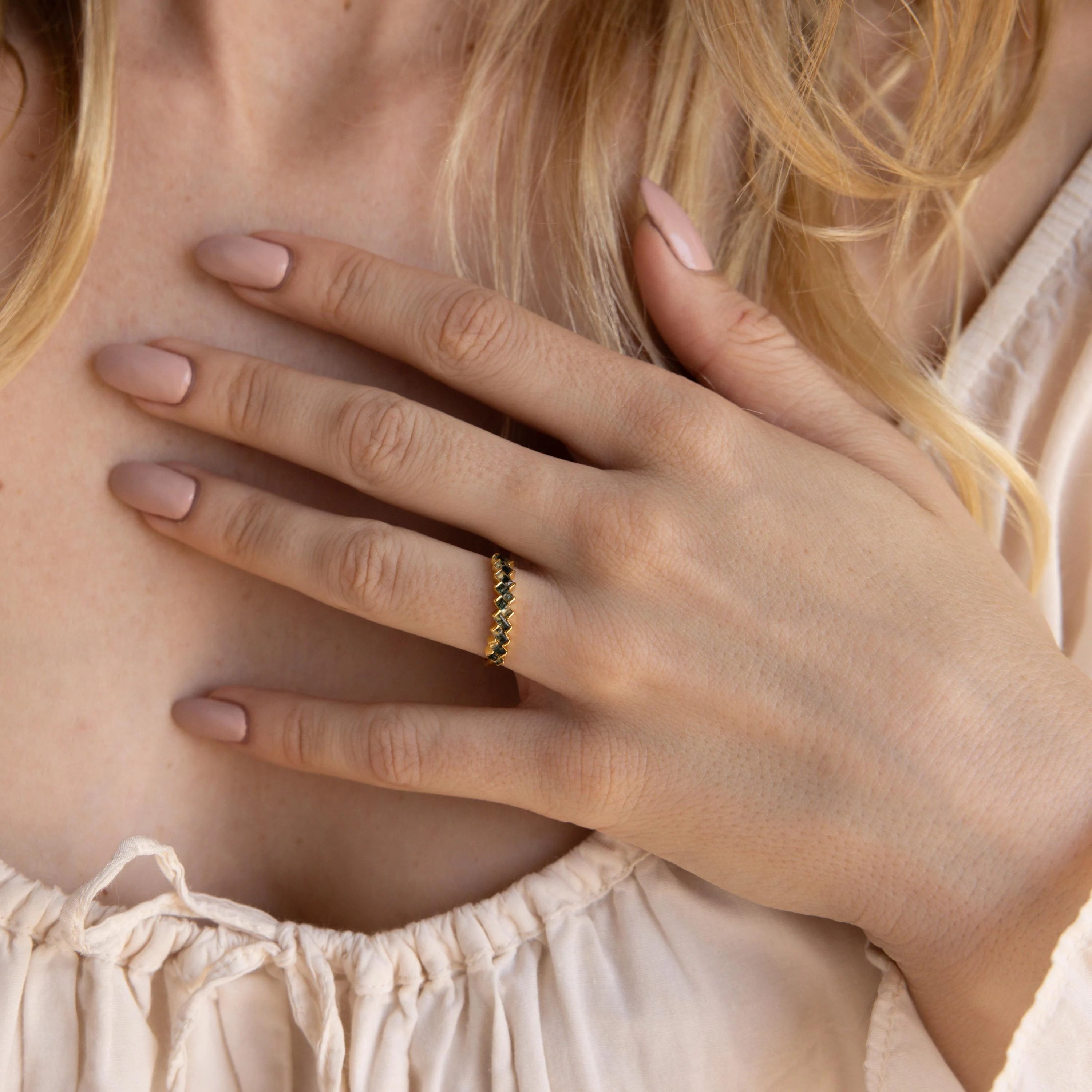 A woman's hand with nude nails rests on her chest, adorned with the Moss Agate Eternity Ring featuring gold and green baguette-cut gemstones.