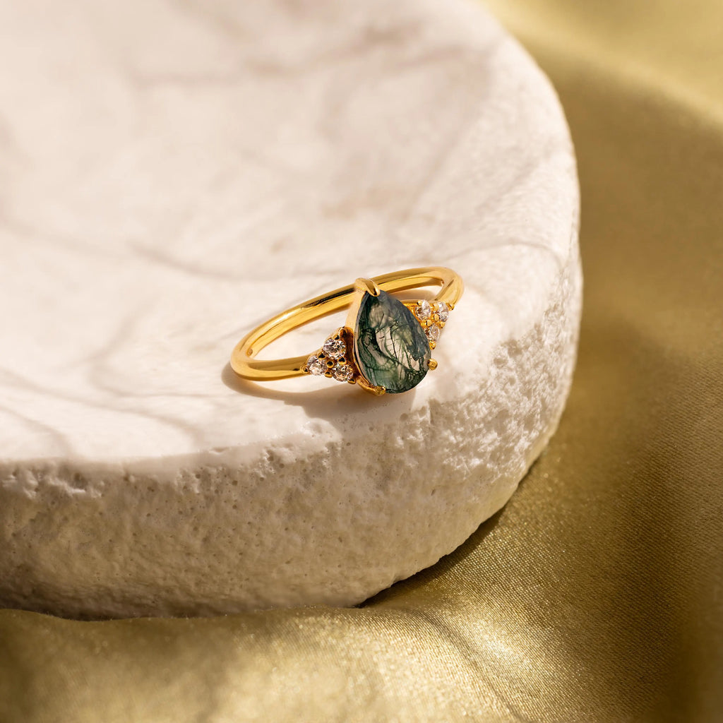 Agate Teardrop Ring in 18K Gold with a green gemstone and small clear stones, shown on a textured white surface with a gold fabric backdrop—a stylish moss agate cocktail ring for any occasion.