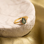 Agate Teardrop Ring in 18K Gold with a green gemstone and small clear stones, shown on a textured white surface with a gold fabric backdrop—a stylish moss agate cocktail ring for any occasion.