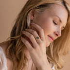 A woman with long blonde hair gently touches her face, displaying the Agate Teardrop Ring in 18K Gold, featuring a striking green gemstone on her finger.