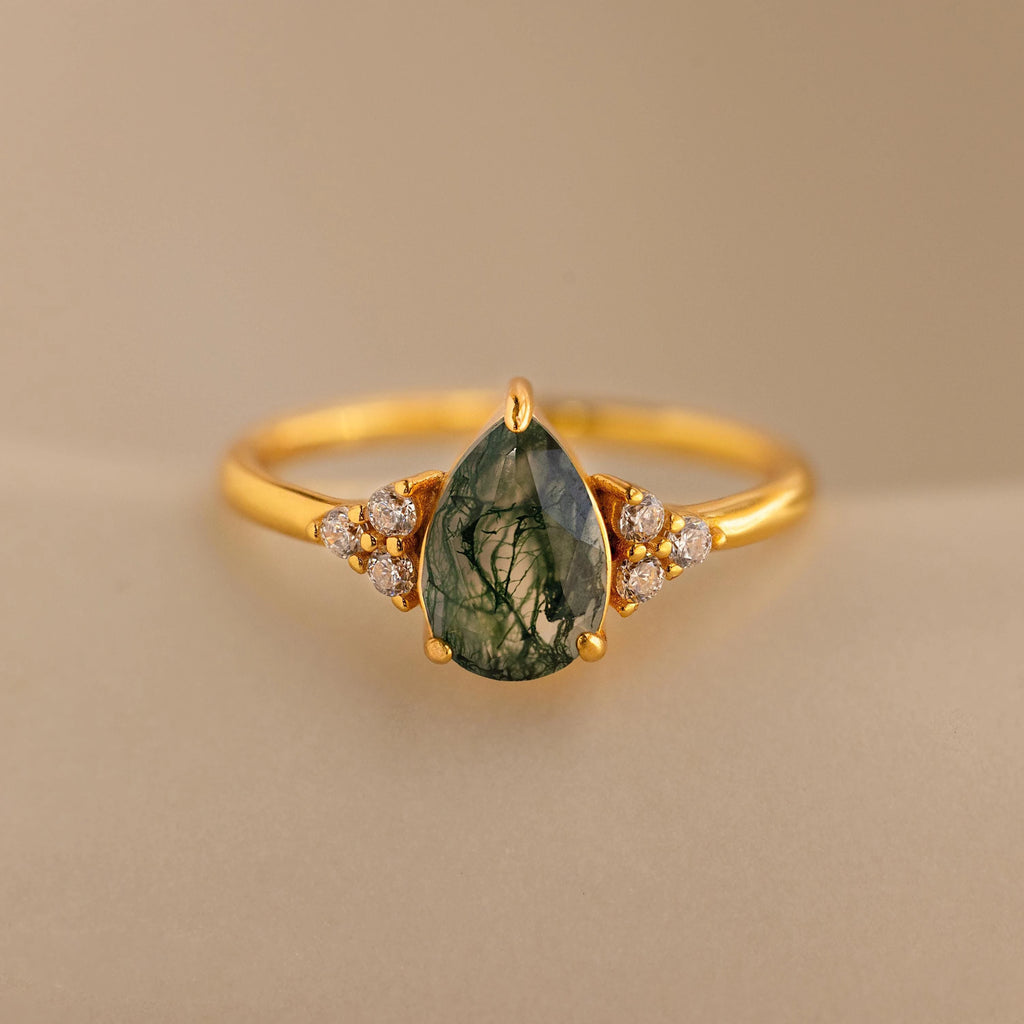 The Agate Teardrop Ring features a green, teardrop-shaped moss agate stone flanked by small white gemstones, showcased on a beige background.