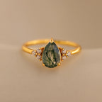 The Agate Teardrop Ring features a green, teardrop-shaped moss agate stone flanked by small white gemstones, showcased on a beige background.