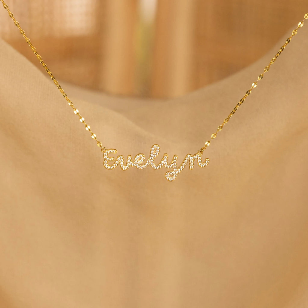The Pave Dream Script Name Necklace, featuring "Evelyn" in cursive pavé letters, shimmers beautifully against a soft beige background.