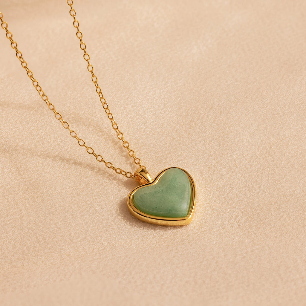 The Aventurine Heart Necklace features a gold chain and a heart-shaped green aventurine pendant, shown on a soft beige background.
