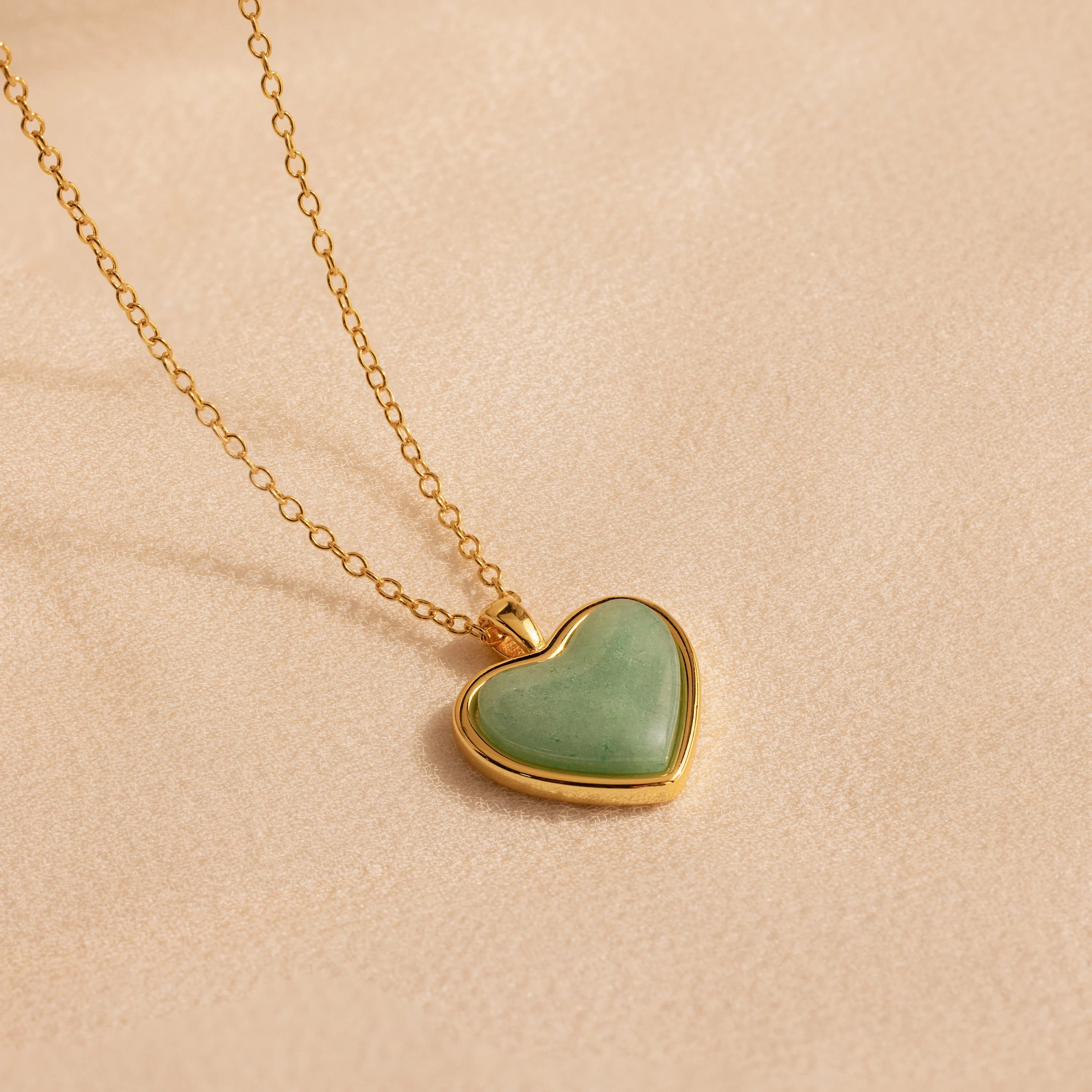 The Aventurine Heart Necklace features a gold chain and a heart-shaped green aventurine pendant, shown on a soft beige background.