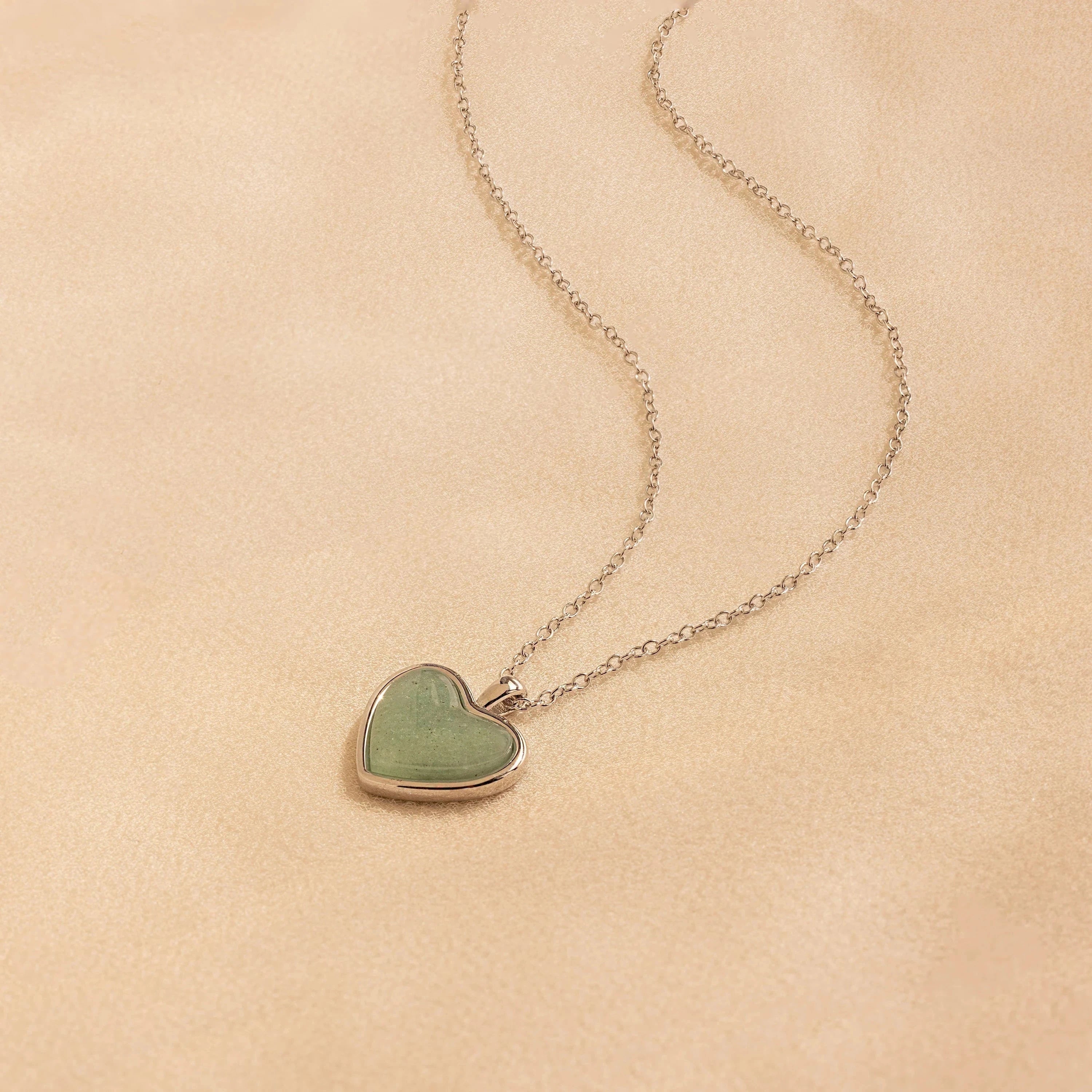 The Aventurine Heart Necklace features a green heart-shaped pendant on a silver chain, elegantly displayed on a beige surface.