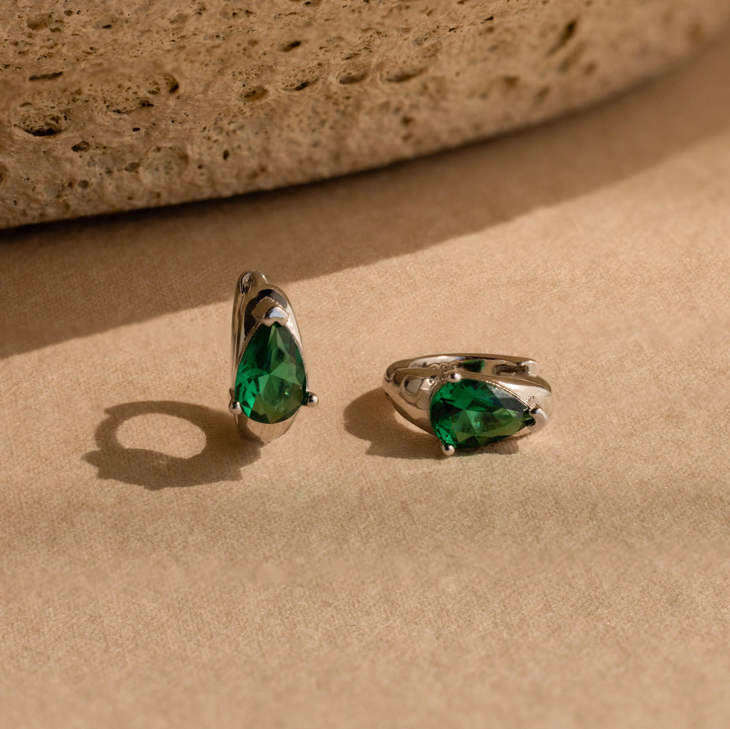 Emerald Teardrop Huggies—silver dainty hoop earrings featuring green, teardrop-shaped gemstones—are displayed on a beige textured surface.