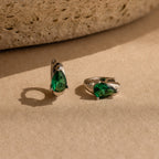Emerald Teardrop Huggies—silver dainty hoop earrings featuring green, teardrop-shaped gemstones—are displayed on a beige textured surface.