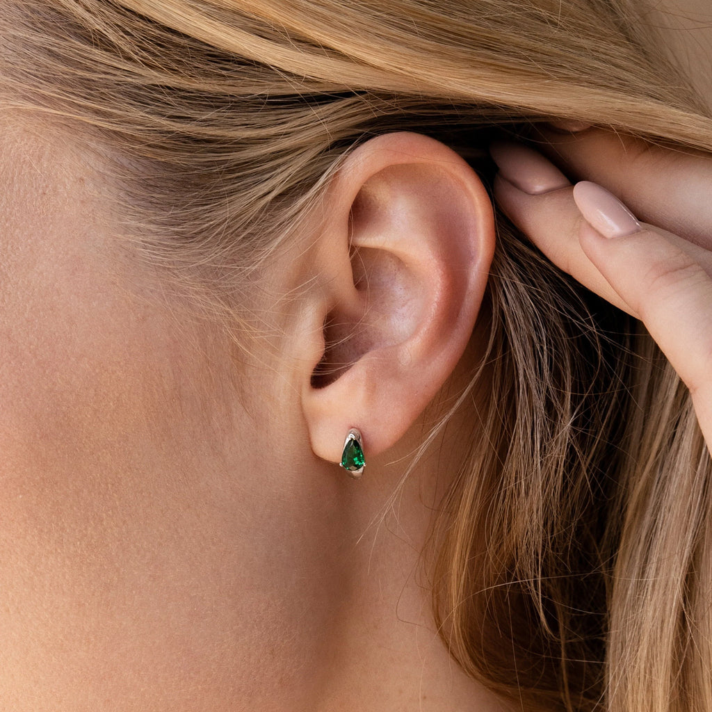 Close-up of a woman's ear wearing Emerald Teardrop Huggies—small green emerald teardrop-shaped earrings.