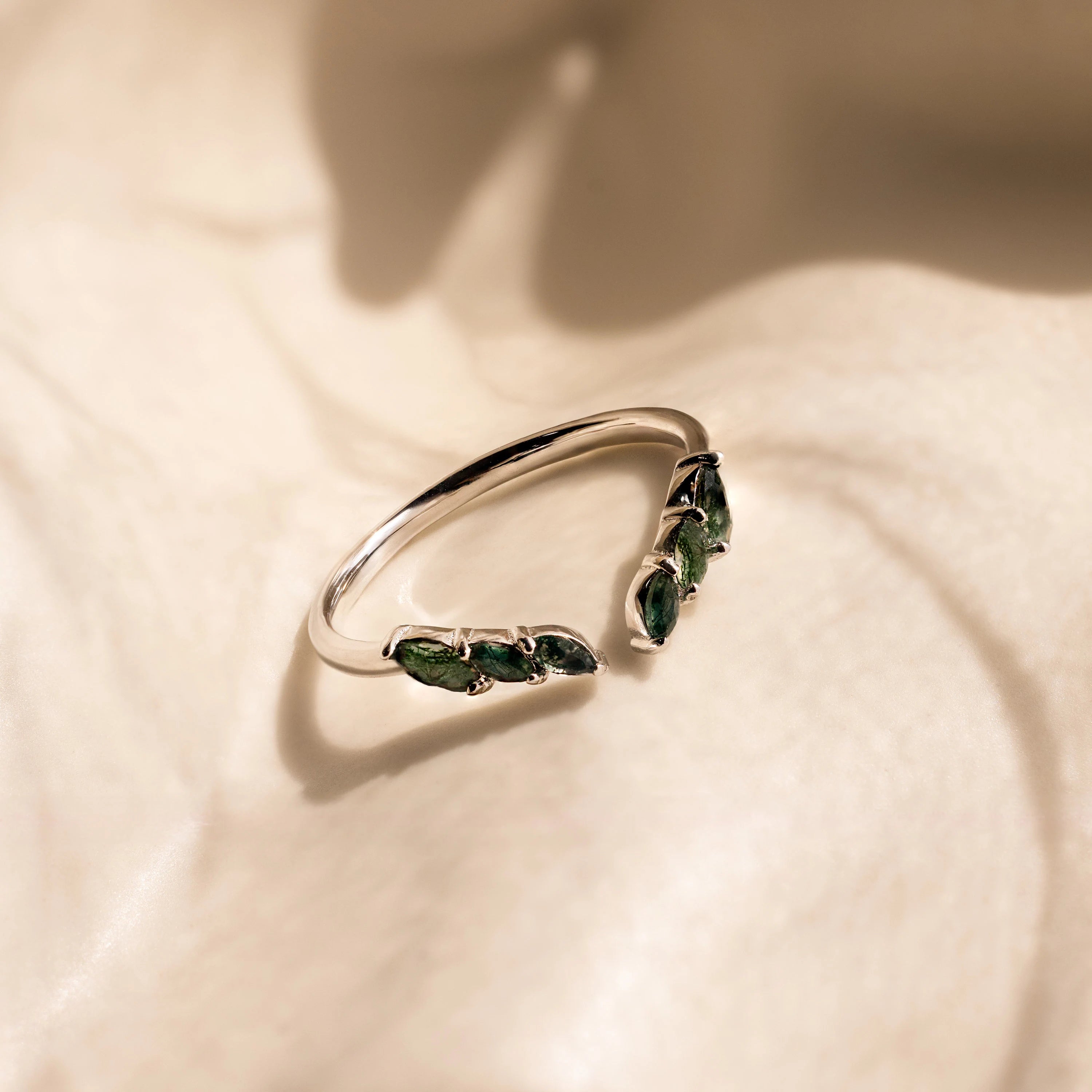 The Open Moss Agate Chevron Ring is a delicate silver piece featuring green moss agate gemstones shaped like leaves, displayed elegantly on a soft beige surface.