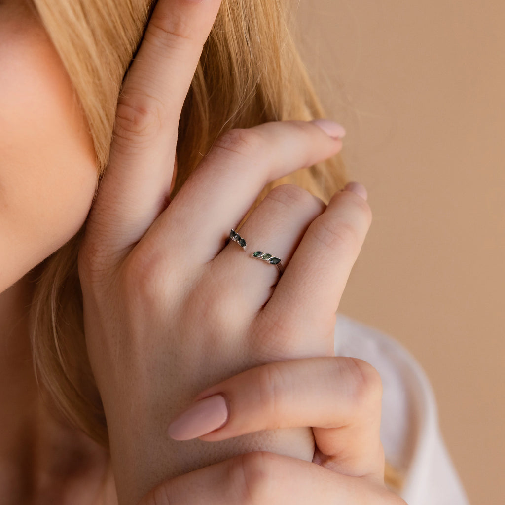 A blonde-haired woman touches her face, highlighting the Open Moss Agate Chevron Ring—a delicate promise ring with green stones—on her finger.