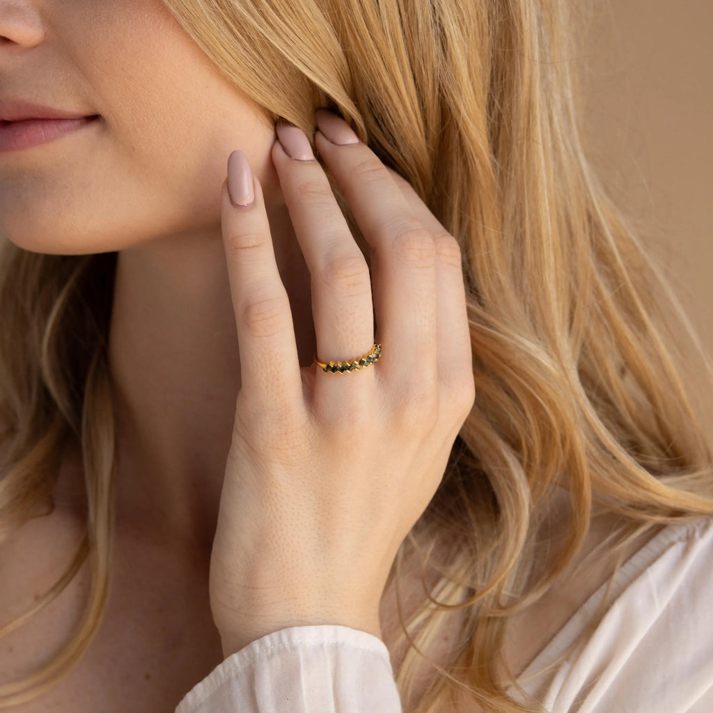 A woman with light hair touches her face, wearing the Moss Agate Eternity Ring—a gold band adorned with sparkling black stones inspired by nature.