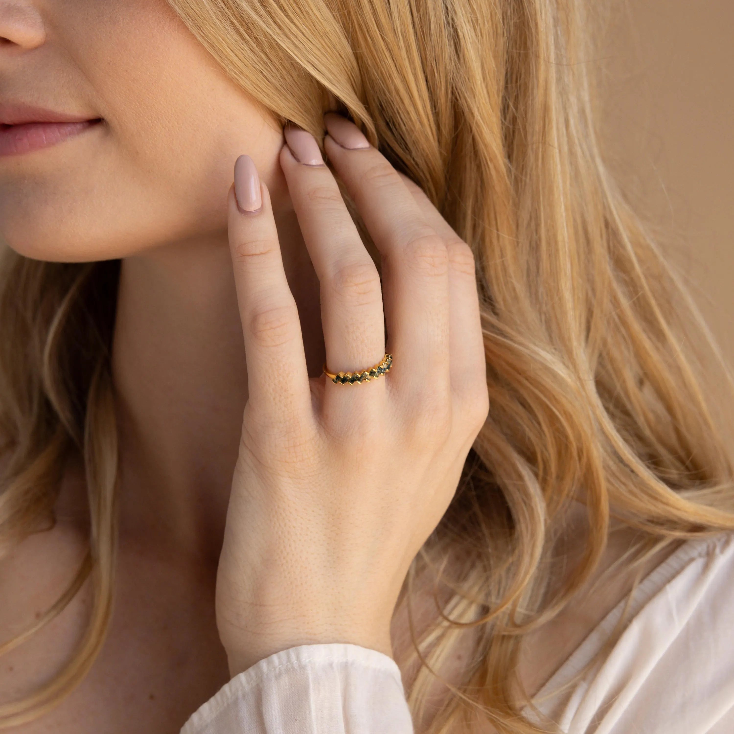 A woman with light hair touches her face, wearing the Moss Agate Eternity Ring—a gold band adorned with sparkling black stones inspired by nature.