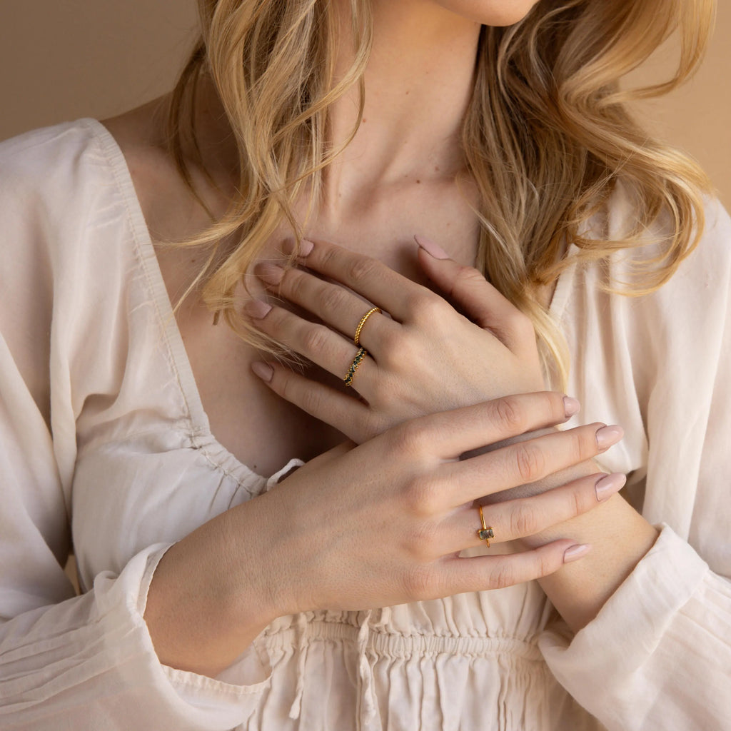 A woman in a cream blouse displays gold rings on her fingers, her hands gently crossed on her chest, highlighting the Moss Agate Eternity Ring.