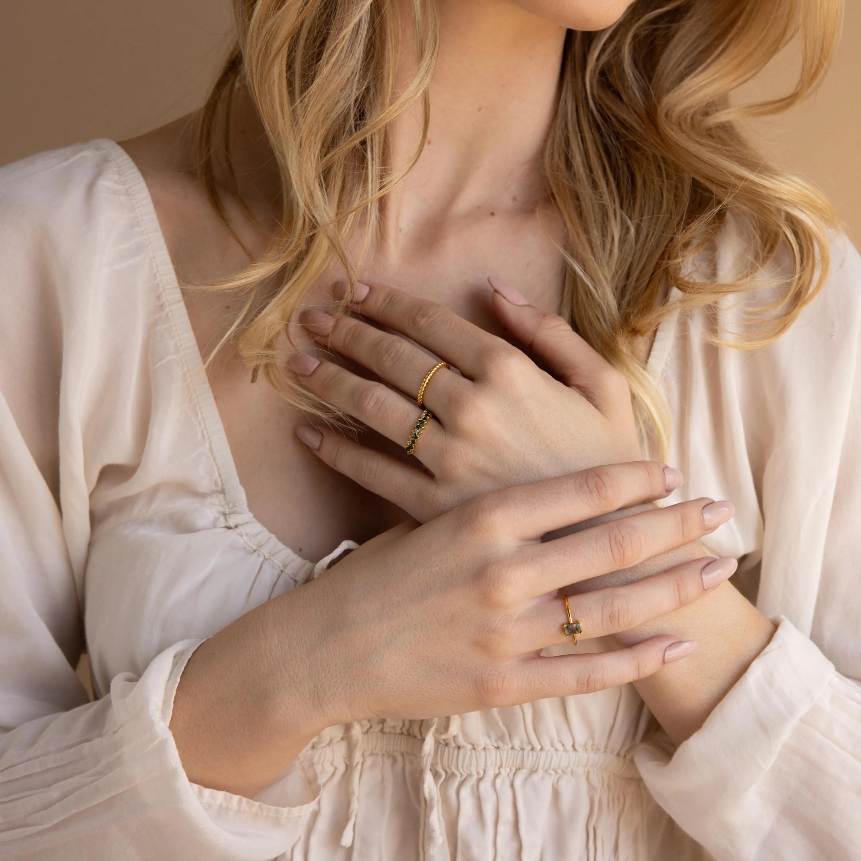 A woman in a cream blouse displays gold rings on her fingers, her hands gently crossed on her chest, highlighting the Moss Agate Eternity Ring.
