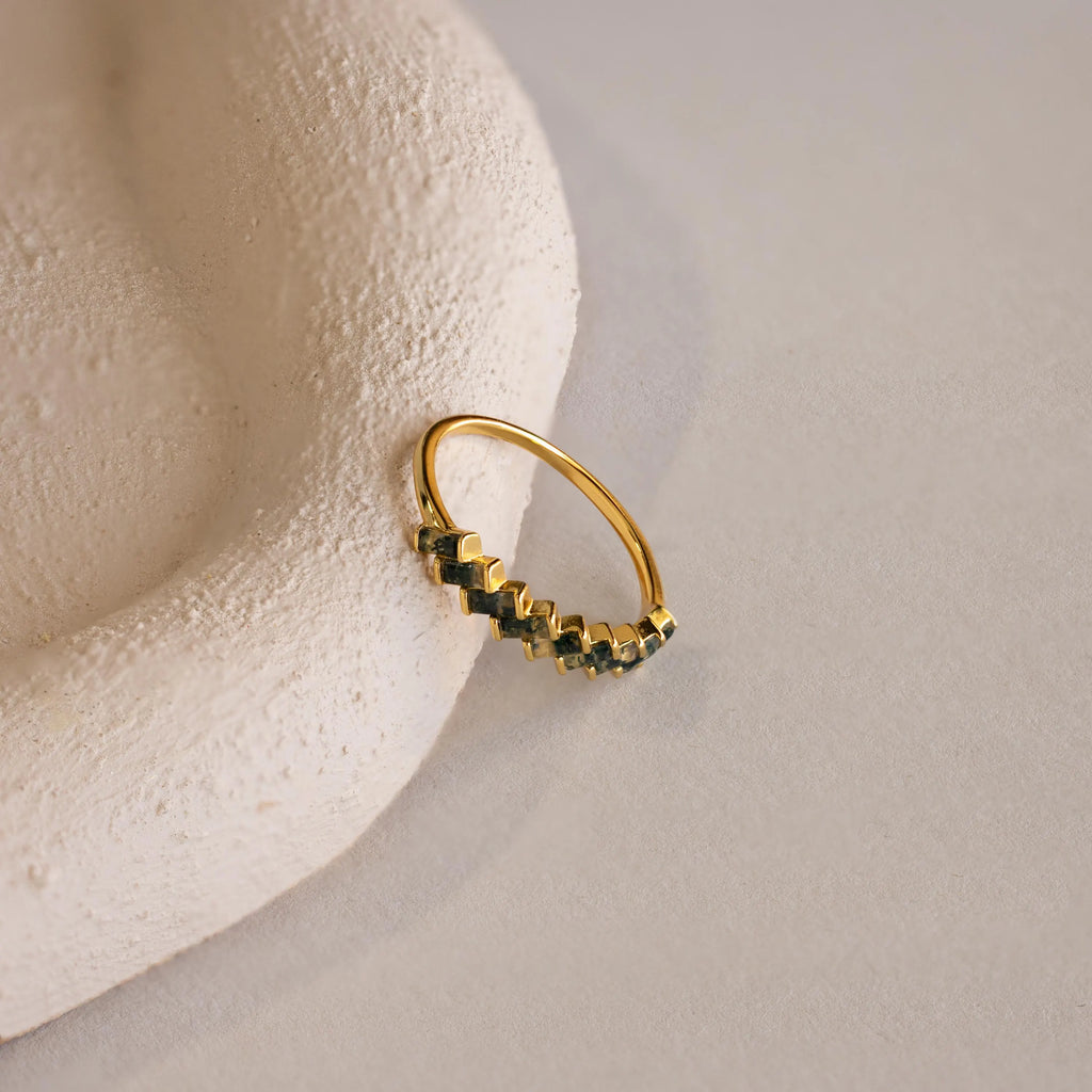 The Moss Agate Eternity Ring features a row of baguette-cut moss agate gemstones, elegantly set on a gold band and displayed on a textured light beige surface.